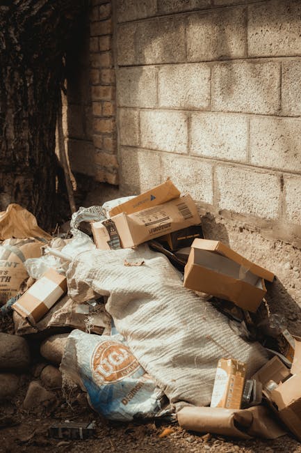 A collection of overflowing rubbish bins and waste accumulated on a paved sidewalk area next to a parking lot, with various types of waste including black and black plastic bags, flattened cardboard b