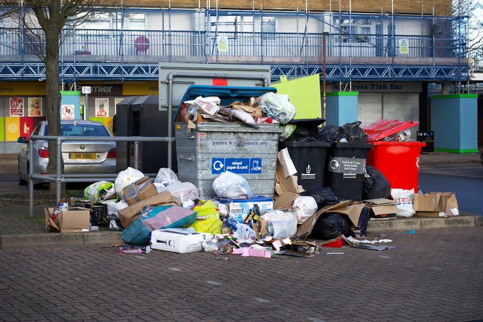 A worker in a high-visibility orange and yellow vest is operating a large, red rubbish collection vehicle on the side of a paved street. The back of the vehicle, which features hydraulic arms and a lo