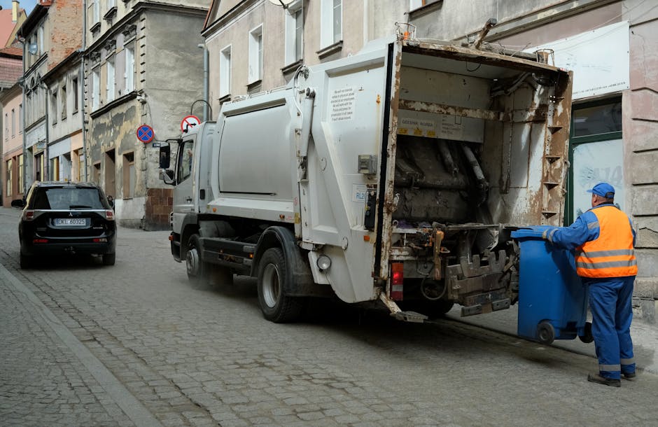 A male waste management worker wearing a blue cap, high-visibility yellow jacket, and bright orange trousers with reflective strips is using a leaf blower or vacuum device to manage litter on a grassy