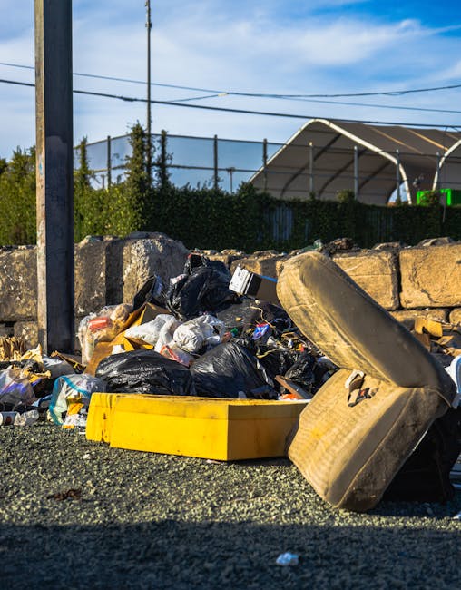 A large collection of mixed waste and rubbish is piled around and atop multiple wheeled bins in an outdoor urban setting. The refuse includes various cardboard boxes, plastic bags, paper and packaging